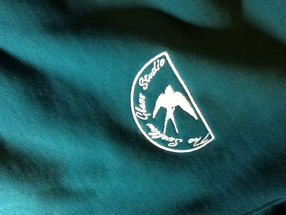 An embroidery design of a stylized heron silhouette within a circular emblem featuring the text "The Glass Studio" above it.