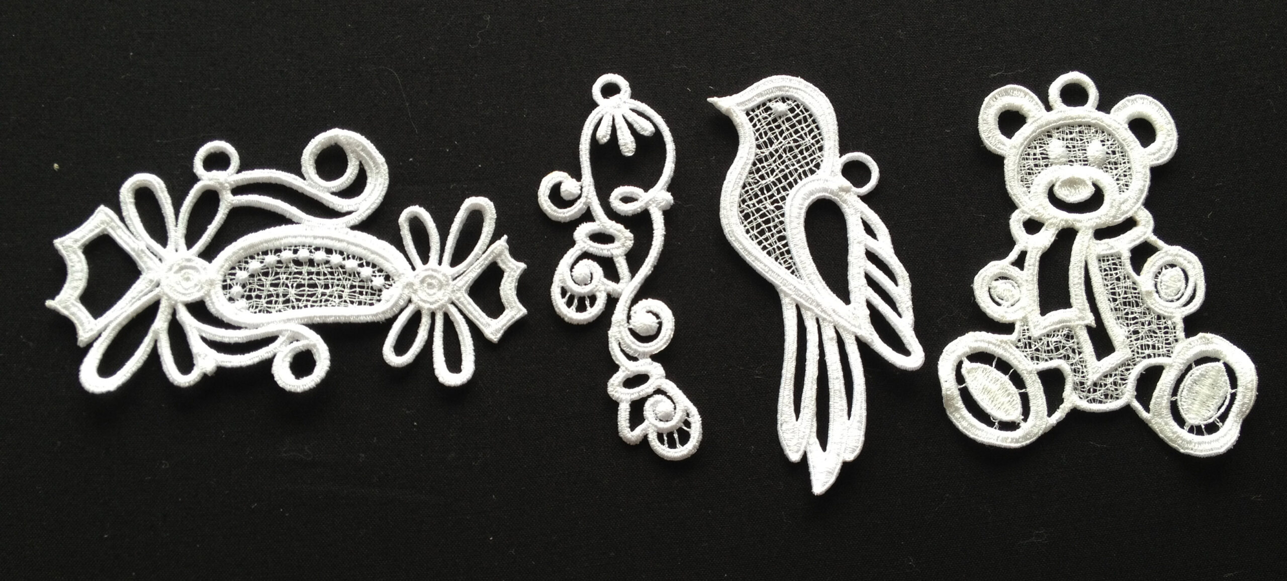 An embroidery design of delicate lace motifs including a decorative bow, a whimsical bird, a curly flourish, and a cute teddy bear.