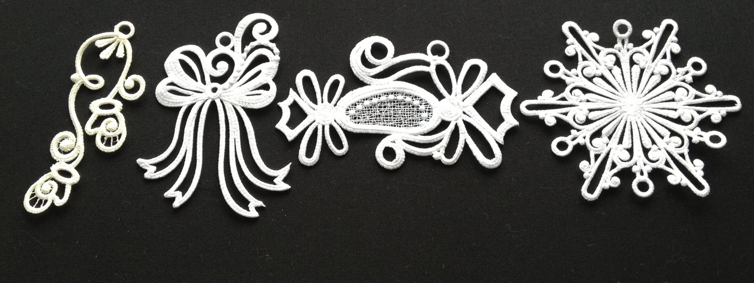 An embroidery design of ornate white motifs including a delicate swirl, a bow, a decorative candy shape, and a snowflake.