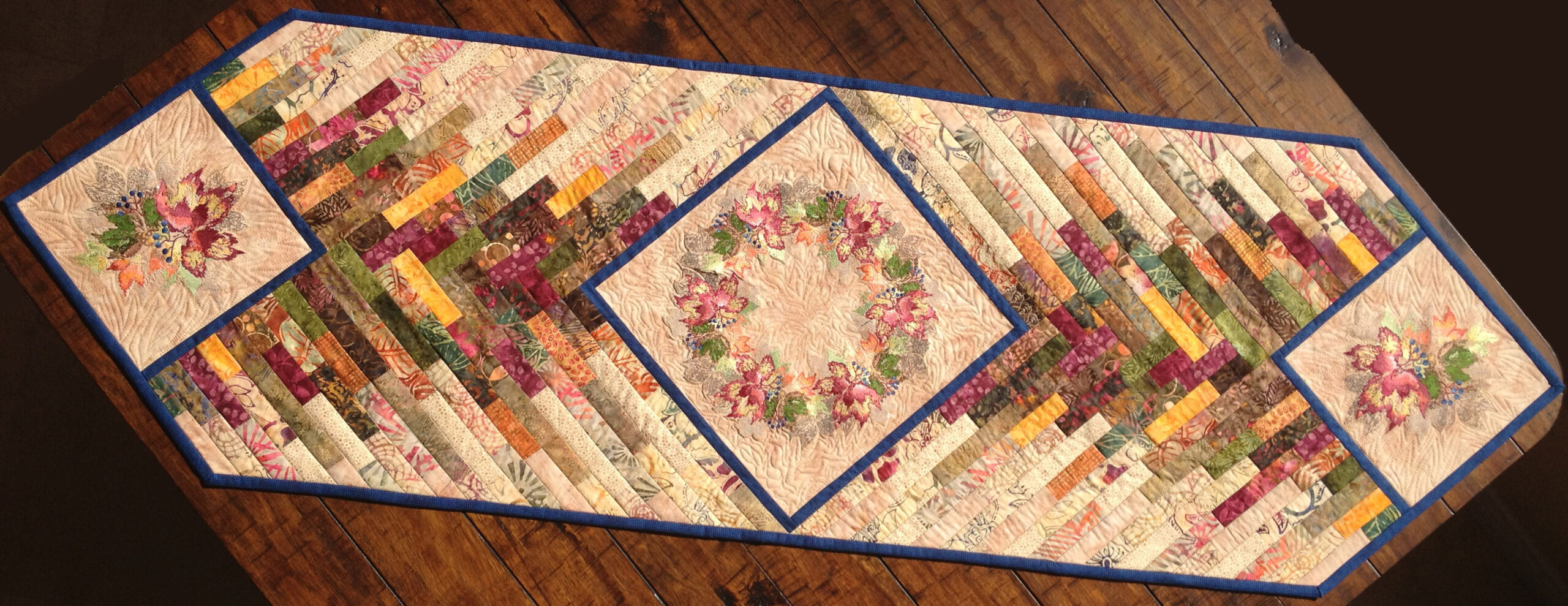 An embroidery design of a floral motif is featured on a patchwork runner with vibrant fabric strips arranged in a geometric pattern.