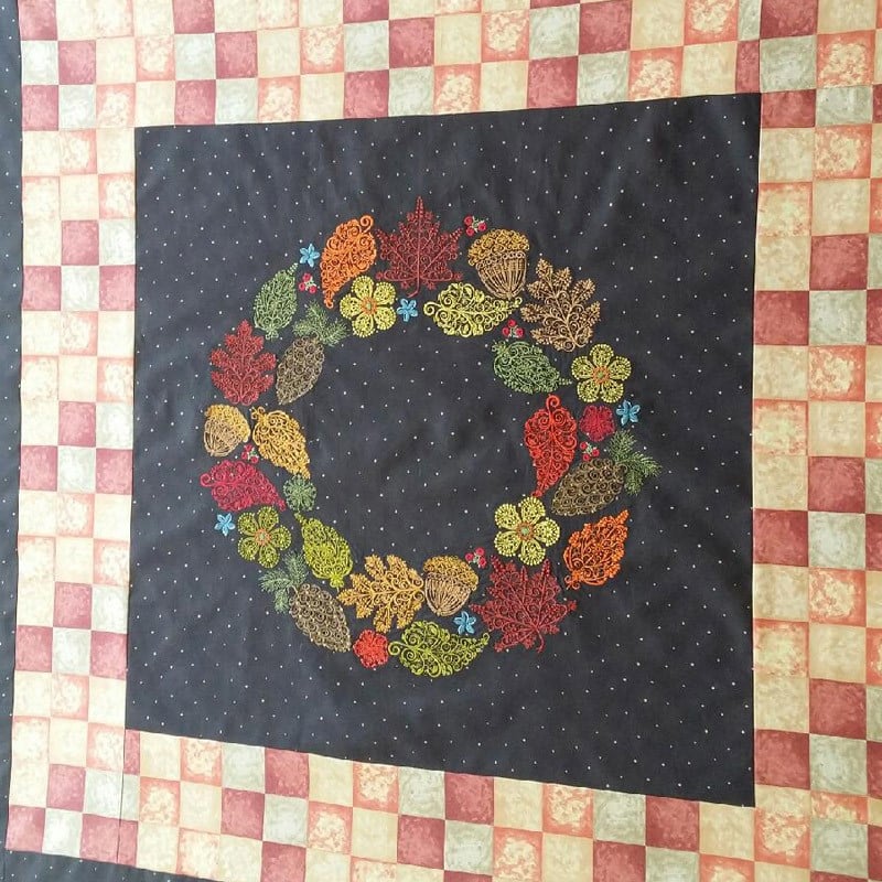 An embroidery design of a vibrant wreath made of colorful leaves, flowers, and acorns intricately stitched against a dark background.