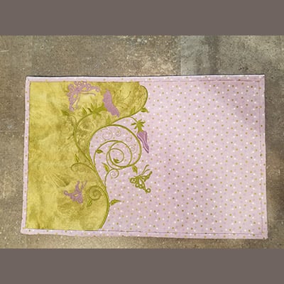 An embroidery design of delicate vine patterns adorned with flowers and butterflies in shades of green and purple on a soft pink background with polka dots.