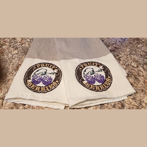 An embroidery design of purple berries within a circular frame featuring the words "FRUIT" and "BERRIES" on a towel.