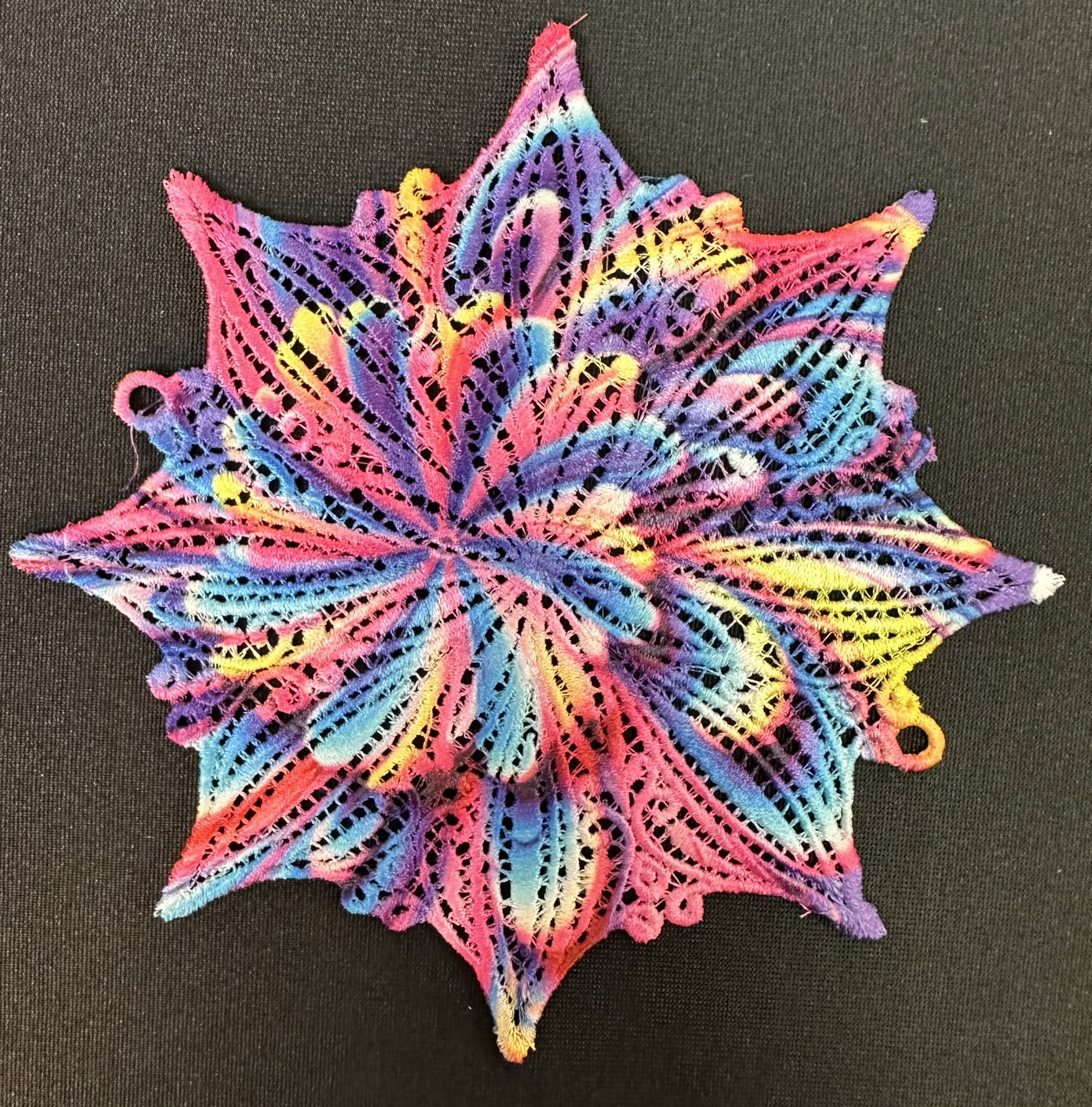 An embroidery design of a vibrant, multilayered lace pattern with a star shape featuring swirls of pink, blue, yellow, and purple.
