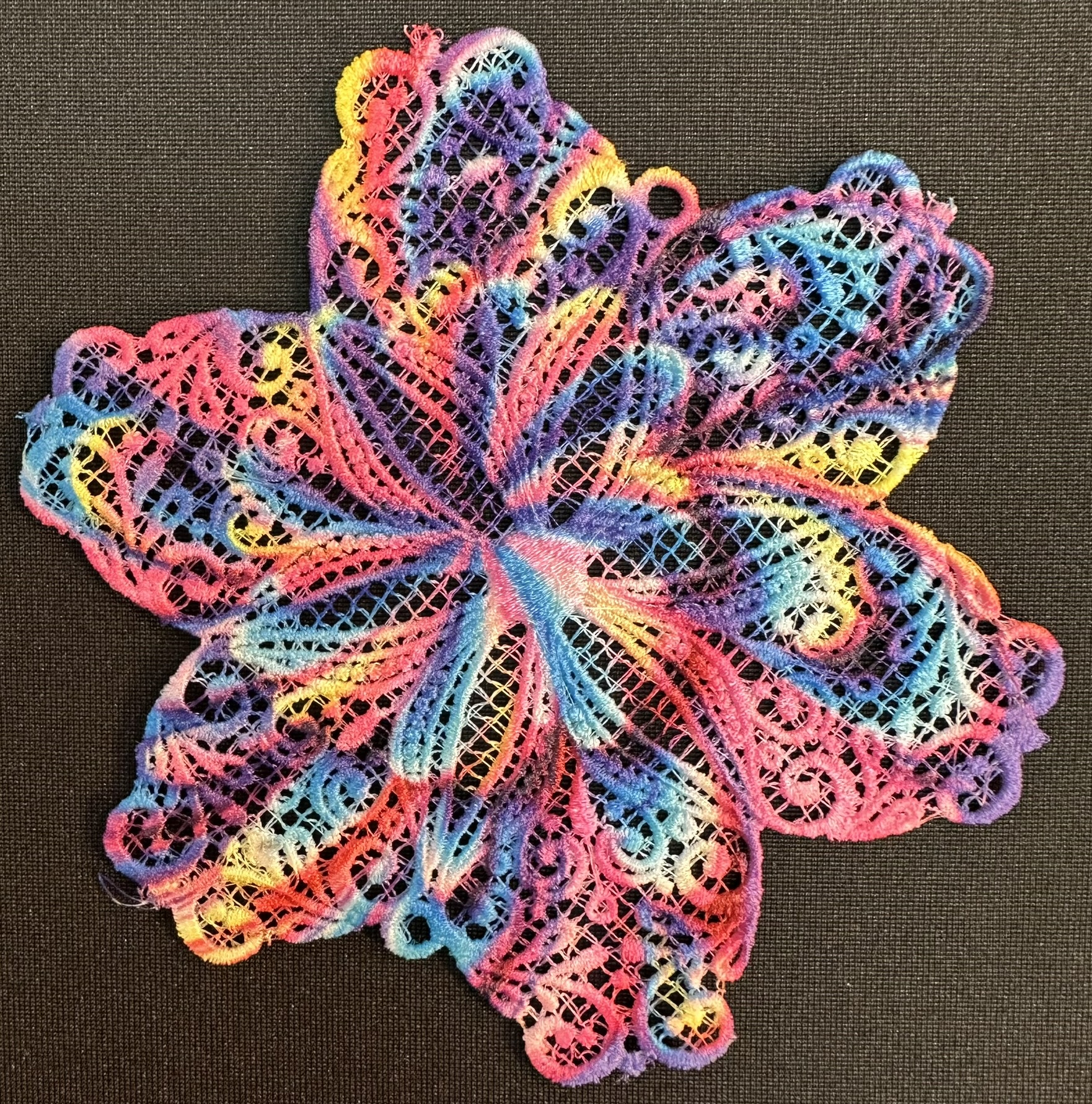 An embroidery design of a colorful, lace-like flower with intricate patterns in shades of pink, blue, and yellow.