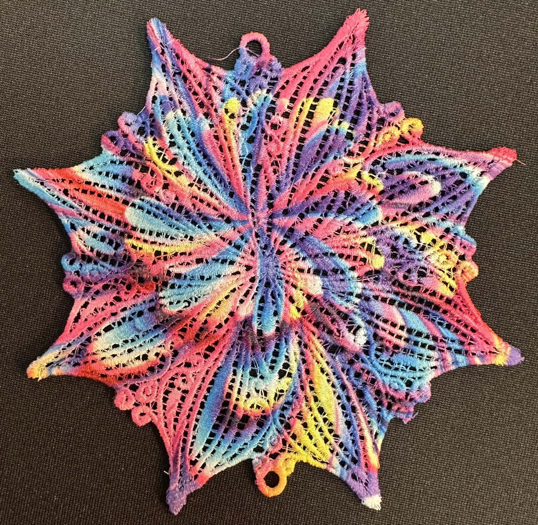 An embroidery design of a vibrant, star-shaped lace pattern featuring a blend of pink, blue, and yellow colors.