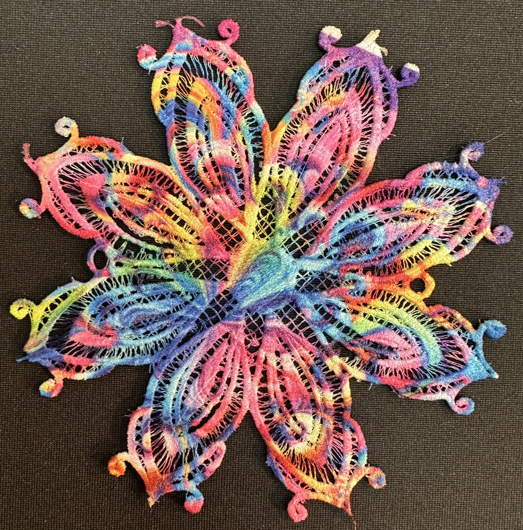 An embroidery design of a colorful, intricately patterned flower with swirling petals in vibrant hues of pink, blue, and yellow.
