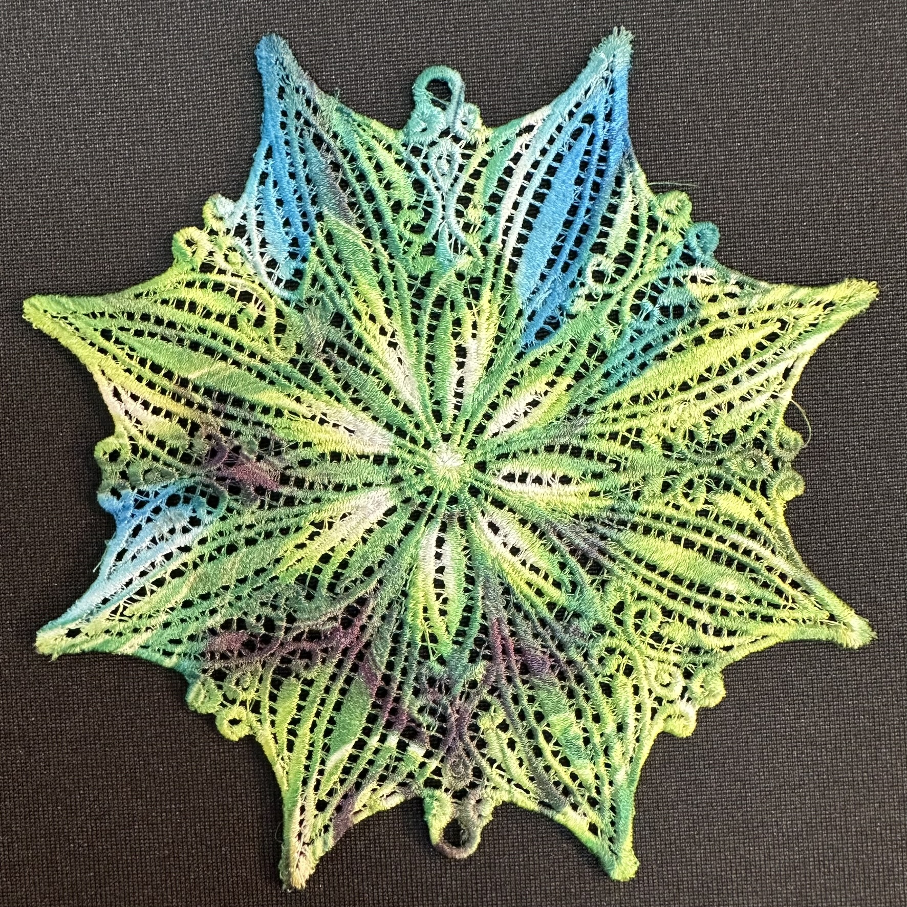 An embroidery design of a vibrant, intricate lace pattern shaped like a snowflake, featuring a blend of green, blue, and subtle purple hues.