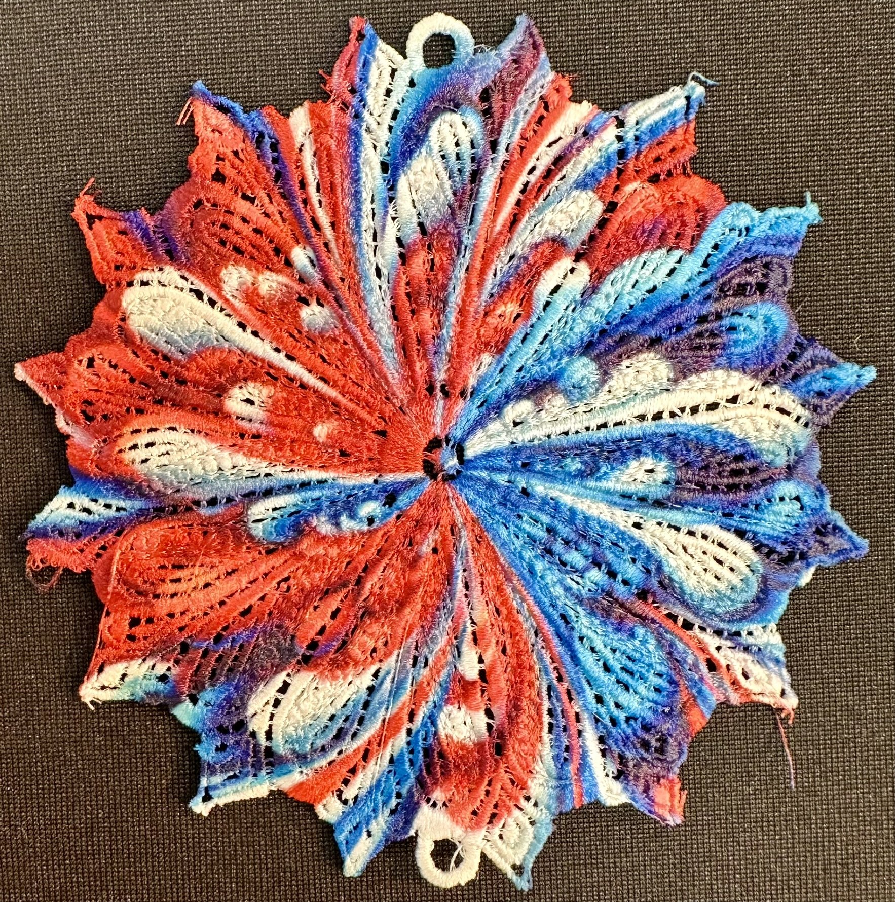An embroidery design of a vibrant, circular pattern featuring swirling shades of red, blue, and white, intricately layered to create a lace-like effect.