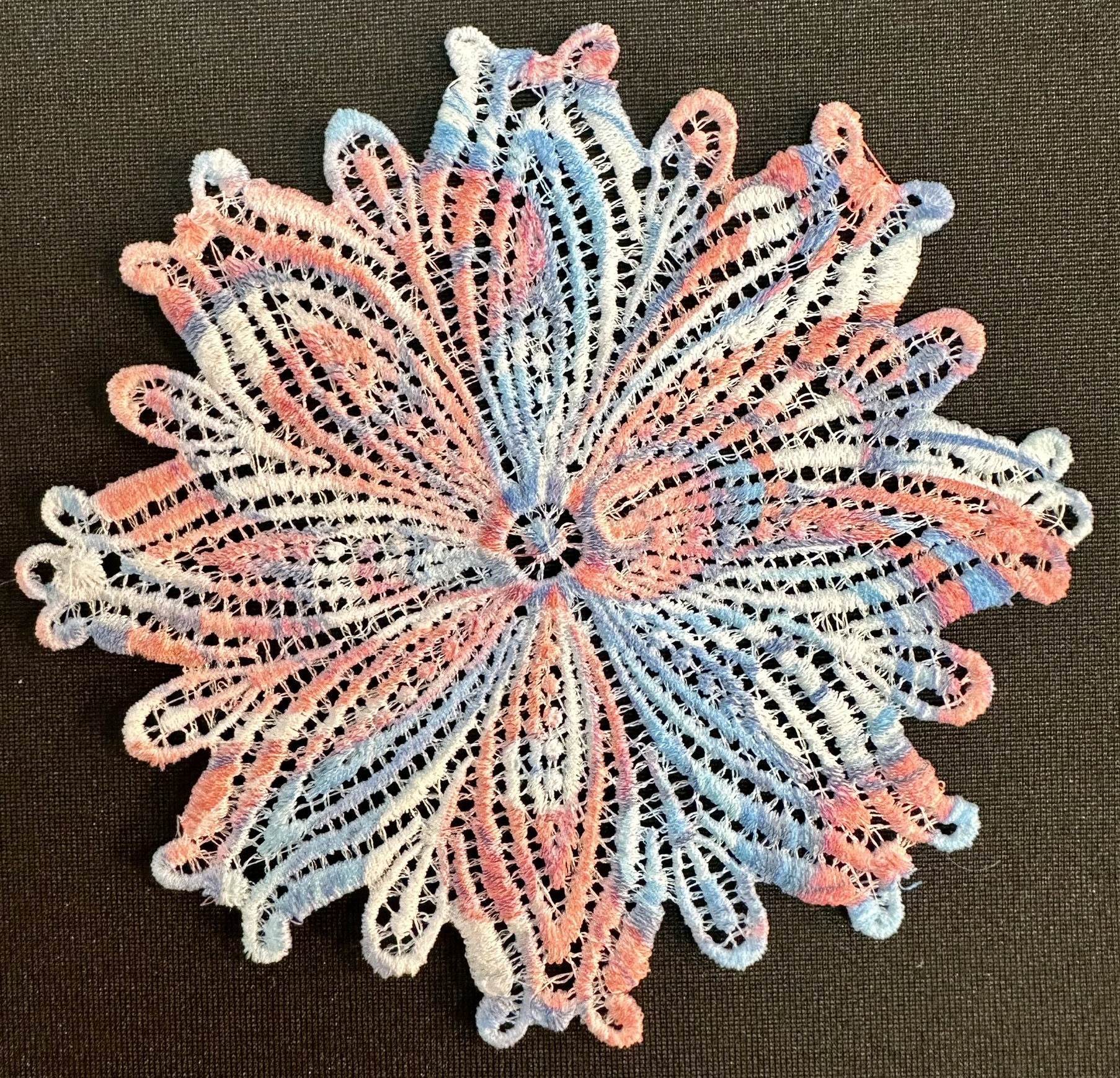 An embroidery design of a delicate, lace-like floral motif featuring intricate loops and swirls in soft shades of pink and blue.