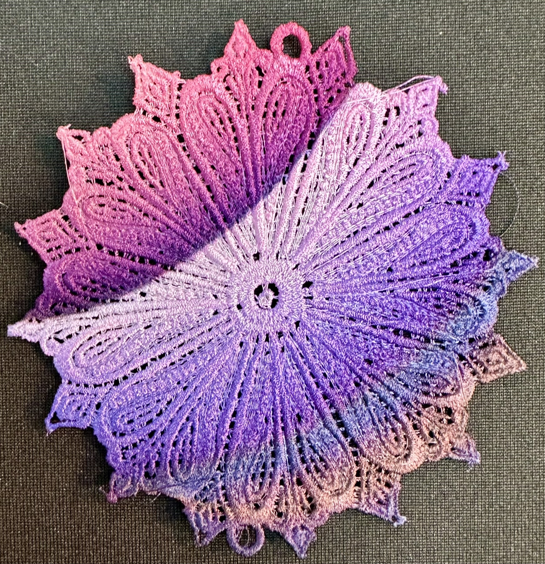 An embroidery design of a circular, multi-layered floral pattern featuring a gradient of purple hues.